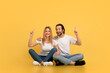 Glad young caucasian male and female showing fingers up to empty space, sitting on floor