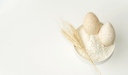 On a light background, a plate with wheat flour, two eggs and wheat ears, ingredients for baking.  Space, copy space for text.  Flat lay, top view.  The concept of minimalism.