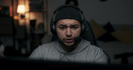 Close-up of guy in black cap playing online game. Man has headphones on head and is talking to the players. Boy plays the final match and loses. He takes off headphones, nervous about defeat.