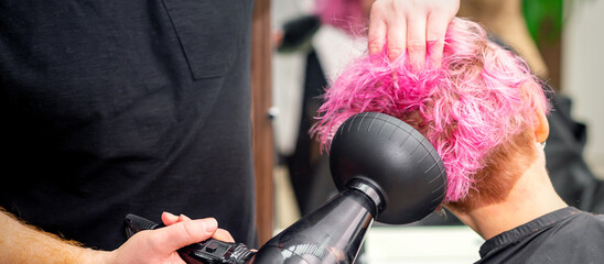 Drying short pink bob hairstyle of a young caucasian woman with a black hair dryer with the brush by hands of a male hairdresser in a hair salon, close up