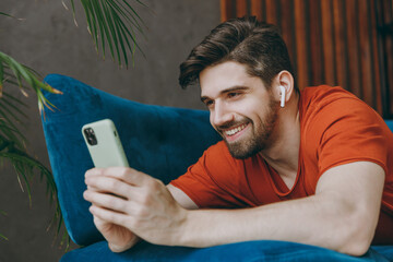 Wall Mural - Young man wears red t-shirt listen music in earphones use mobile cell phone lay down on blue sofa stay at home flat rest relax spend free time in living room indoors grey wall. People lounge concept.