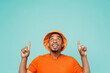Young happy man of African American ethnicity 20s wear orange t-shirt hat point index finger overhead indicate on workspace area copy space mock up isolated on plain pastel light blue cyan background.