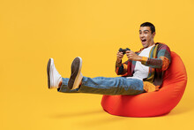 Full Body Young Middle Eastern Man 20s He Wear Casual Shirt White T-shirt Sit In Bag Chair Hold In Hand Play Pc Game With Joystick Console Isolated On Plain Yellow Background People Lifestyle Concept.