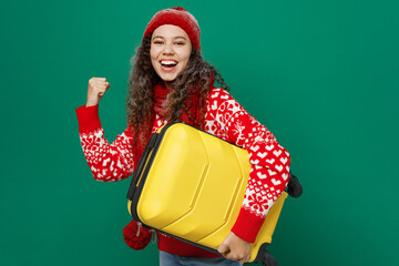 Wall Mural - Traveler woman wear red cozy sweater hat hold suitcase do winner gesture isolated on plain dark green background. Tourist travel abroad in free spare time rest getaway Air flight trip journey concept.