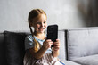 © F8  \ Suport Ukraine - Portrait of cute little cute girl holding smartphone and resting at sofa at home. Choosing favorite music or cartoons,texting messages, browsing internet, watching video, playing games onphone