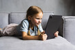 © F8  \ Suport Ukraine - My favorite pastime. Smiling small child girl sit on comfy sofa in modern studio apartment hold tablet computer read book online use handy web app. Little kid rest on cozy couch play videogames on pad