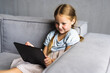 © F8  \ Suport Ukraine - Smiling little girl lying on couch, enjoying playing online game on digital tablet computer. Addicted to technology, happy small kid using funny applications, web surfing information.