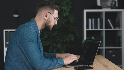 Young bearded coder writing code using laptop, focused man coding in dark office room, application software creation