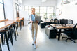 © Kateryna - Modern trendy confident successful young mixed race woman, wearing stylish business clothes, go across the creative office, holding an open laptop in her hands, looking at the camera, smiling