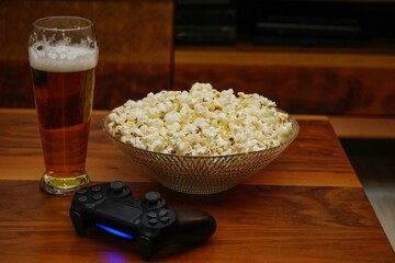 Glass of beer, a bowl of popcorn, and a video game controller on top of a wooden table