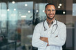 © Liubomir - Portrait of young happy and smiling doctor, man in medical coat and stethoscope smiling and looking at camera, Arab doctor with crossed arms working inside modern clinic office.