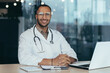 © Liubomir - African american doctor portrait, man working inside modern clinic office at table using laptop, doctor in medical coat and stethoscope smiling and looking at camera.