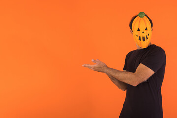 Person with pumpkin mask celebrating Halloween, pointing to the side with fingers, on orange background. Concept of celebration, All Souls' Day and All Saints' Day.