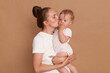 © sementsova321 - Portrait of female with bun hairstyle in white t shirt standing with her infant child, mother kissing her baby daughter, expressing gentle, posing isolated over brown background.
