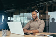© Liubomir - Young office worker working inside office building, using laptop to watch video and headphones, businessman on break.