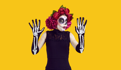 Young woman gets very scared on Halloween or Day of the Dead. Fearful lady in Catrina costume isolated on yellow background does protective gesture and looks at camera with frightened face expression