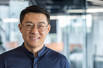 Close up photo portrait of successful young Asian investor, man in glasses smiling and looking at camera in shirt, businessman working inside modern office building, satisfied accountant financier.