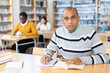 © JackF - Concentrated adult hispanic man spending time in library, reading books and making notes. Self-education concept