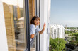 Woman looks out the window and waving hand in residential district in the city, urban top view from the outside, sunny weather. Concept of living in apartment building, quarantine and loneliness