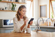 © Nina Lawrenson/peopleimages.com - Senior woman reading phone news, mobile apps and social media notification in morning home kitchen. Relax retirement lady typing smartphone, online network website and 5g tech connection in apartment