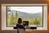 Woman works on laptop while sitting by the table in front of panoramic window with great view on mountains. Remote work and escaping to nature concept