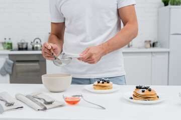 Canvas Print - Cropped view of man pouring powdered sugar in sieve near honey and pancakes in kitchen.