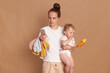 © sementsova321 - Indoor shot of serious young adult woman with bun hairstyle wearing white t shirt standing with her toddler daughter and using mobile phone with concentrated expression, isolated over brown background