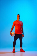 Full-length portrait of male volleyball player posing with ball isolated on blue studio background in neon light. Sport, gym, team sport, challenges