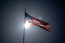 The United States Of The American Flag Blowing In The Wind Of A Bright Sunny Day With The Sun Behind It