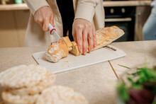 Close Up Of Hands Of A Young Woman Cooks In Her Home Kitchen, Cuts A Baguette Bread With A Knife