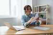 © SHOTPRIME STUDIO - Attentive smiling happy female student in headphones learning from online lectures at home writes down synopsis on sheet. Happy office employee is at video conference using laptop. Copy space