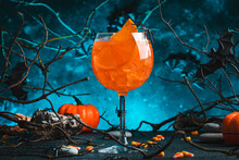Halloween Aperol Spritz Cocktail On Scary Dark Blue Background With Twisted Branches, Bats, Stones, Pumpkin Guards, Corn And Spiders,  Festive Drink For Party