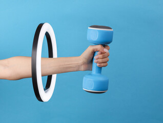 Wall Mural - Woman's hand holds plastic dumbbell through led ring lamp on blue background. Creative idea. Fitness, sport concept