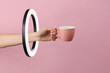 Woman's hand holds empty ceramic cup through led ring lamp on pink background. Creative idea