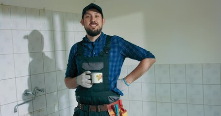 Wall Mural - A busy laborer working on the renovation of a single-family house construction. Guy in work overalls baseball cap relaxes looks out the window sips coffee to wake up. Looking at camera.