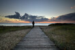 Ein Weg aus Holz führt zum Strand, wo eine Person steht und sich den Sonnenaufgang über dem Meer ansieht