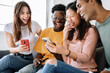 Young group of diverse friends sitting on sofa using mobile phone - Smiling multiracial teenage people laughing having fun together watching funny social media video content on smart phone at home