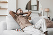 Satisfied handsome young man relaxing on sofa at home in living room, resting after a hard day work, closed eyes, put hands behind head, smiles happily. Relaxation, self care, enjoy life concept