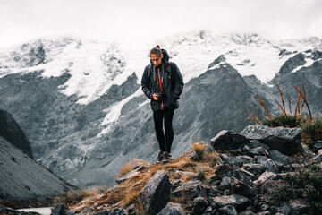 young caucasian man warm in a jacket comfortable sneakers and narrow black trousers with a hanging camera looking at the ground pensively near snowy mountains and gray sky in a nature landscape, mount