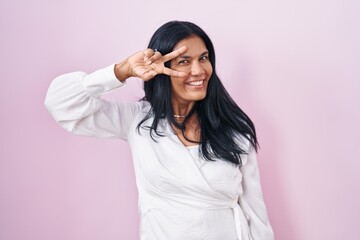 Sticker - Mature hispanic woman standing over pink background doing peace symbol with fingers over face, smiling cheerful showing victory