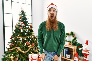 Canvas Print - Redhead man with long beard wearing christmas hat by christmas tree afraid and shocked with surprise and amazed expression, fear and excited face.