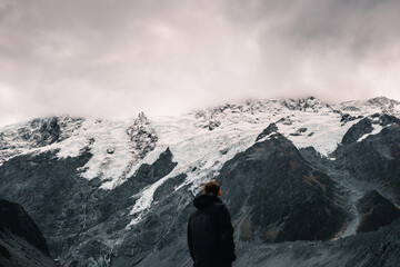 caucasian boy contemplating and admiring the mighty mountains of rock and snow under a gray sky with many clouds on a stormy day, mount cook, new zealand