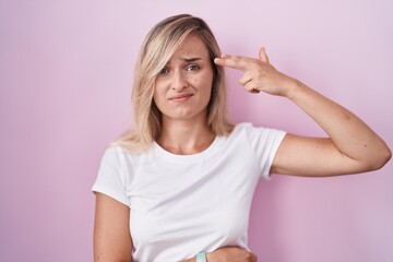 Canvas Print - Young blonde woman standing over pink background shooting and killing oneself pointing hand and fingers to head like gun, suicide gesture.