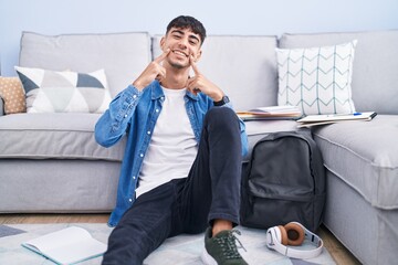 Poster - Young hispanic man sitting on the floor studying for university smiling with open mouth, fingers pointing and forcing cheerful smile