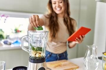 Young beautiful hispanic woman preparing smoothie with blender looking at recipe on tablet at the kitchen