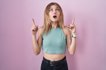 Sticker - Blonde caucasian woman standing over pink background amazed and surprised looking up and pointing with fingers and raised arms.