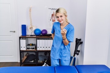 Wall Mural - Young physiotherapist woman working at pain recovery clinic beckoning come here gesture with hand inviting welcoming happy and smiling