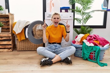 Wall Mural - Young blonde woman doing laundry sitting by washing machine smiling amazed and surprised and pointing up with fingers and raised arms.