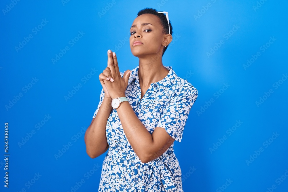 African american woman standing over blue background holding symbolic gun with hand gesture, playing killing shooting weapons, angry face
