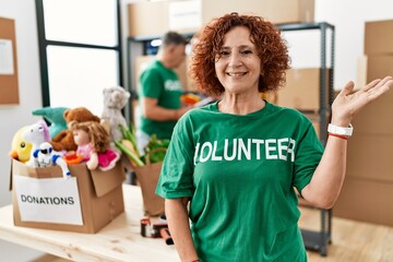 Wall Mural - Middle age woman wearing volunteer t shirt at donations stand smiling cheerful presenting and pointing with palm of hand looking at the camera.